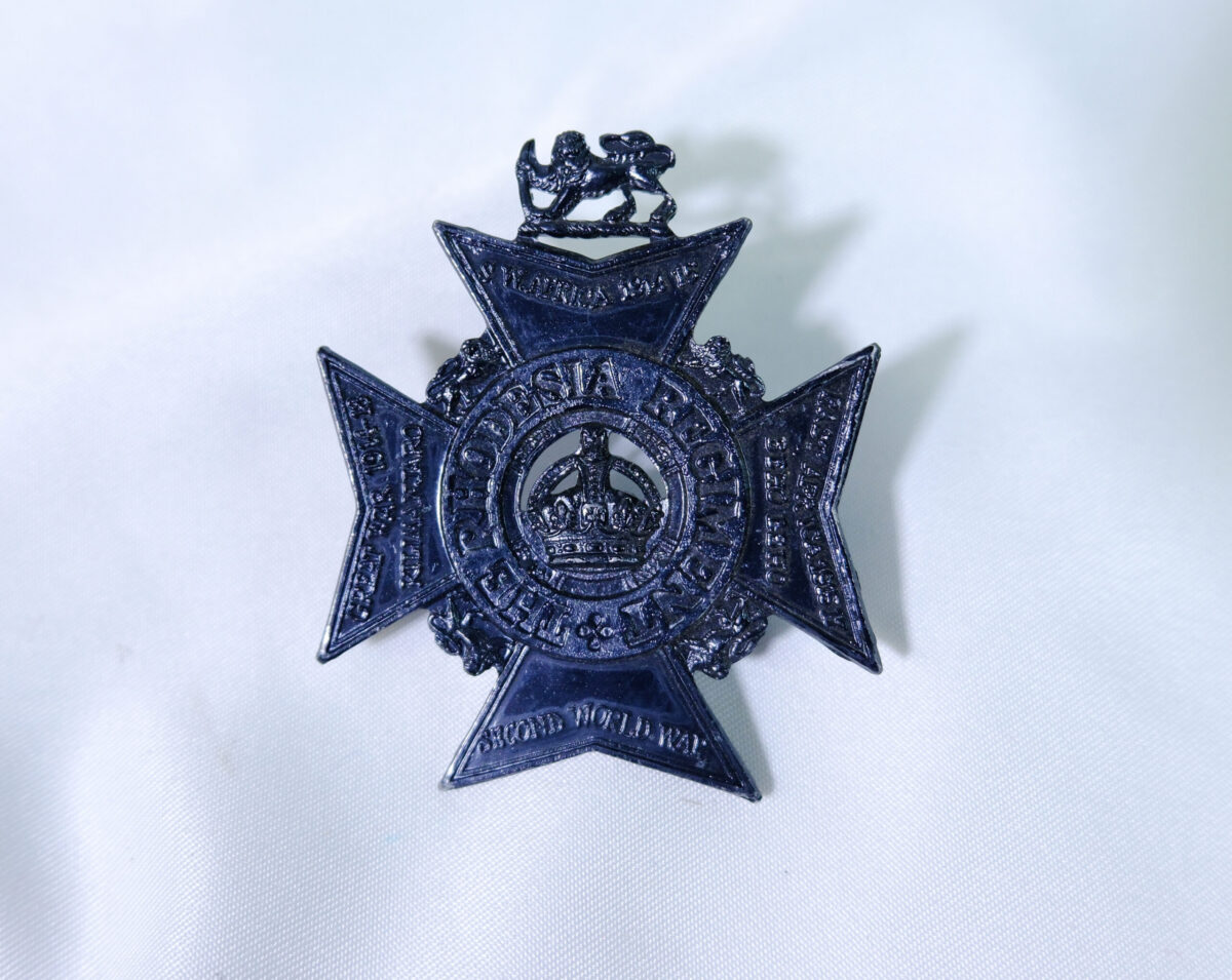 The image shows a dark-toned metal cap badge for The Rhodesia Regiment, featuring a cross design with a central crown encircled by the regiment's name and campaign honours. The image shows a dark-toned metal cap badge for The Rhodesia Regiment, featuring a cross design with a central crown encircled by the regiment's name and campaign honours.